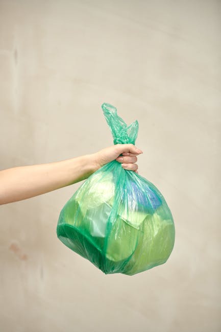 A person's arm extends into the frame from the left, holding a green and blue plastic rubbish bag with a knotted top. The bag appears to be filled with waste and is slightly translucent, revealing som